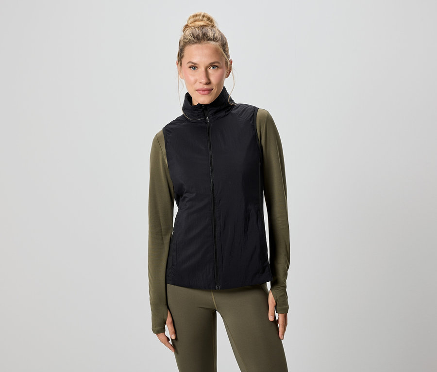 Žena s blond vlasy má na sobě černou vestu přes zelené tričko s dlouhým rukávem a legíny.