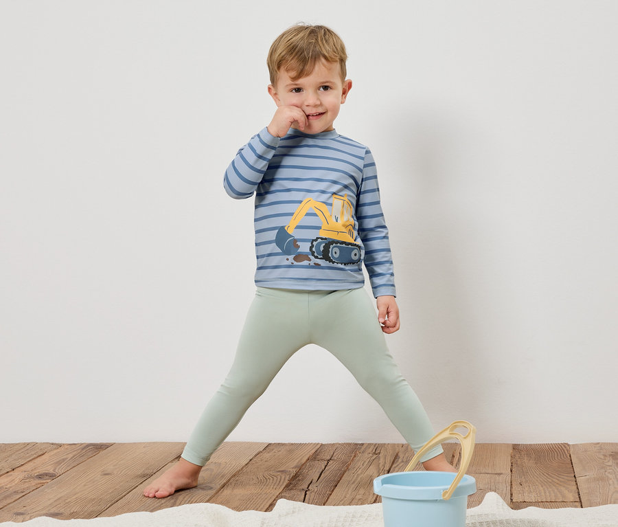 Chlapec stojí bosý na dřevěné podlaze, má na sobě pruhované tričko s dlouhým rukávem s bagrem a světle zelené legíny. Před ním stojí modrý kbelík.