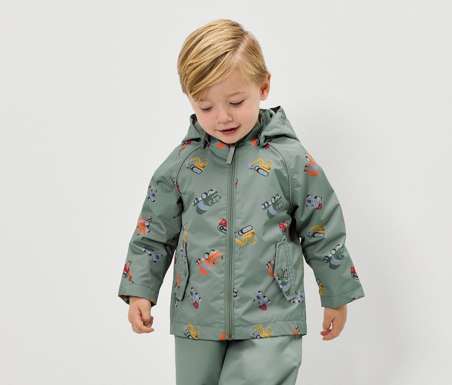 Blonďatý chlapec má na sobě zelenou bundu do deště s motivem bagru a odpovídající kalhoty do deště.