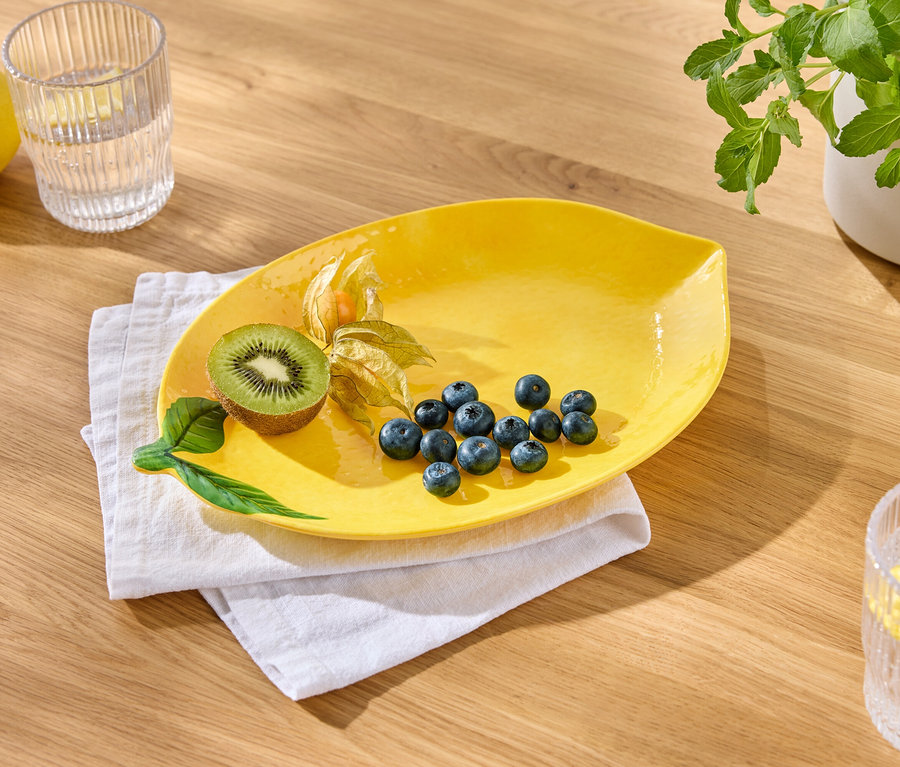 Žlutý talíř s borůvkami, kiwi a mochyní leží na bílém ubrousku. Vedle stojí sklenice vody s citronem a citron.