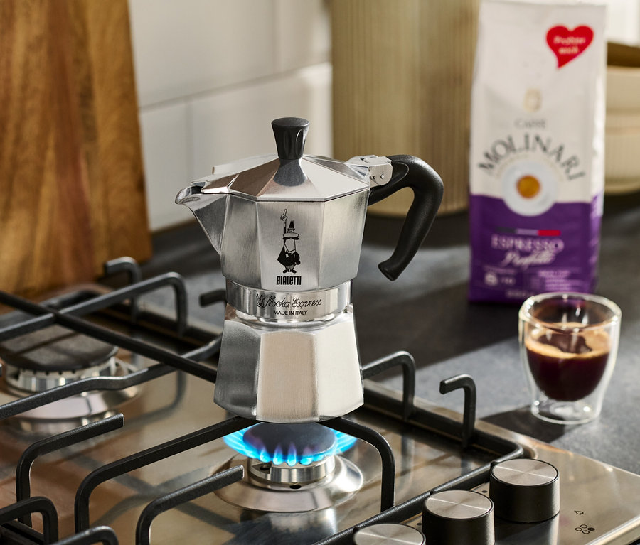 Stříbrný espresso kávovar stojí na plynovém sporáku s modrým plamenem, vedle něj šálek espressa a balíček kávy Molinari.
