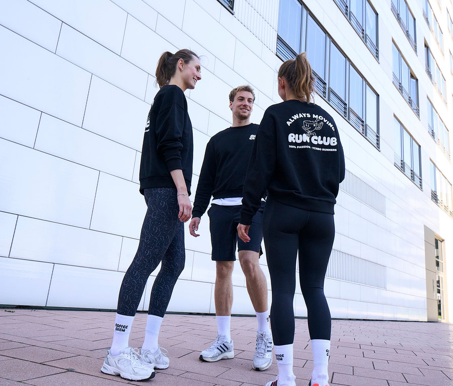 Tři lidé, dvě ženy a jeden muž, stojí usměvaví a povídají si. Všichni mají na sobě černé mikiny a bílé unisex tenisové ponožky Coffee Crew. Muž má černé sportovní šortky. Jedna žena má šedo-černé sportovní legíny »ActivePower«, druhá černé sportovní legíny »ActivePower«.