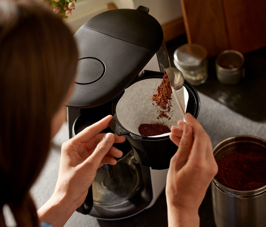 Žena sype kávu lžící do bílého filtru kávovaru »Let's Brew«.