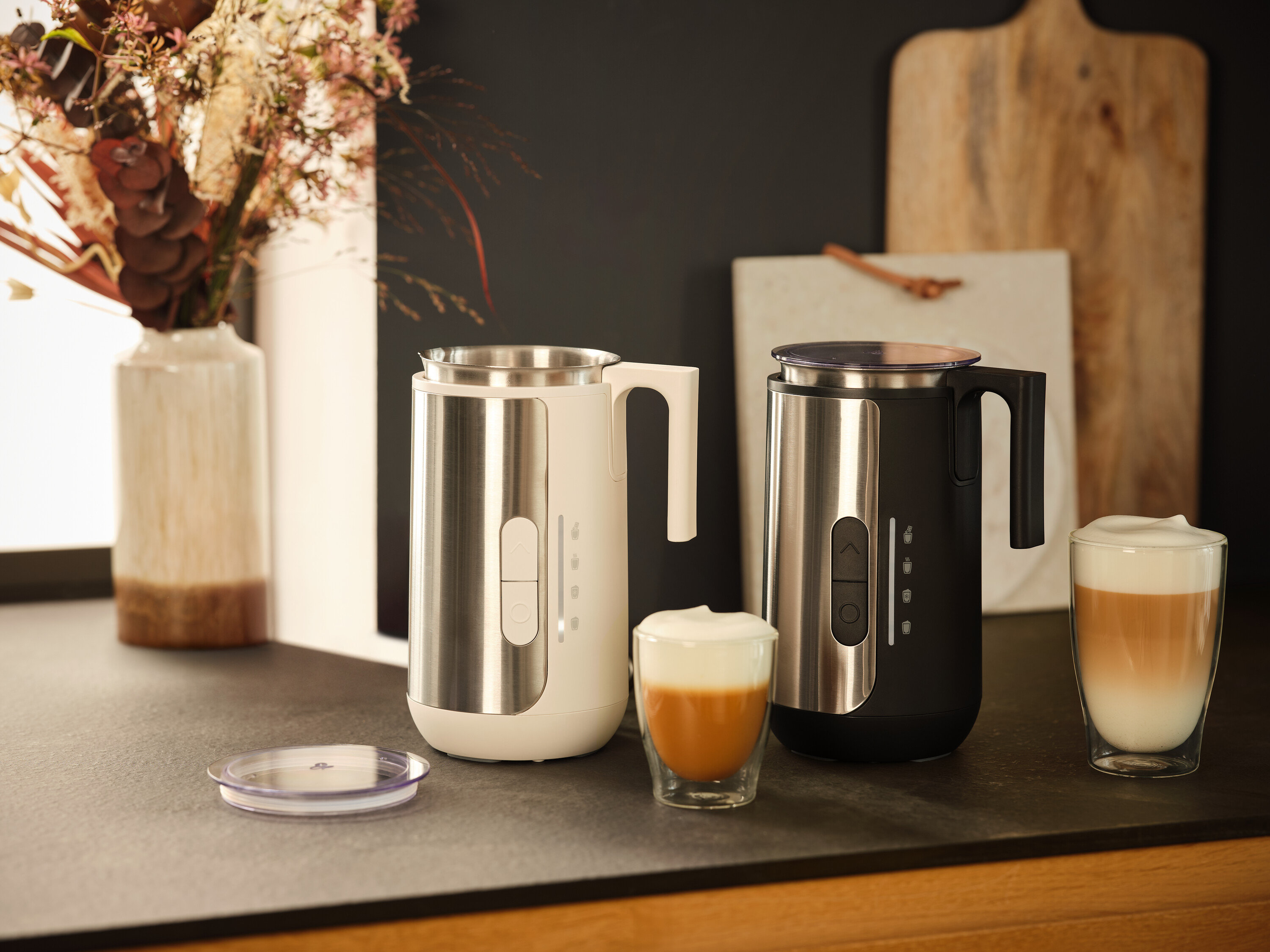 Dva napěňovače mléka, jeden bílý a jeden černý, stojí na desce s dvěma sklenicemi mléčné pěny.