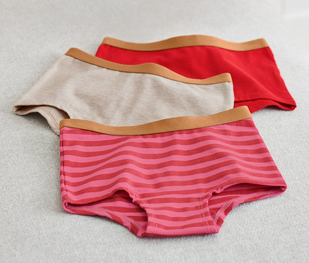 Na světle šedém povrchu leží tři dětské kalhotky, jedny béžové, jedny červené a jedny růžové s proužky. Vlevo je růžová deka.