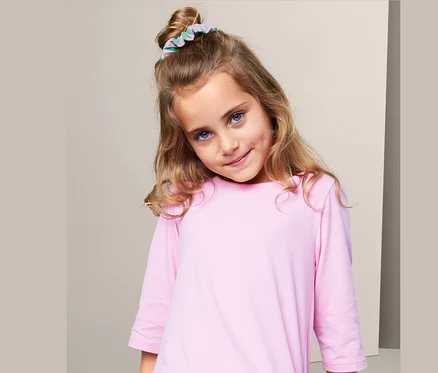 Dívka s blond vlasy a modrýma očima má ve vlasech gumičku do vlasů (3 ks) a na sobě růžové šaty.