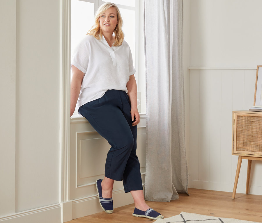 Žena sedí na parapetu v bílé halence ve stylu oversize, ležérních kalhotách chino a espadrilkách.