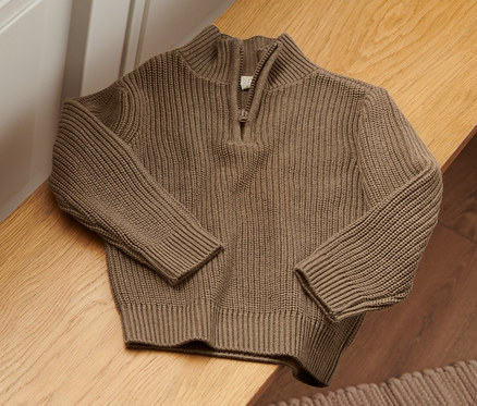 Hnědý svetr troyer leží na dřevěné lavici vedle hračky a fotbalového míče.