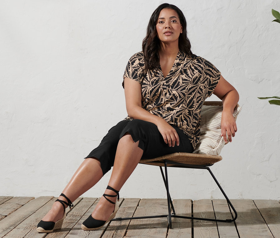 Žena sedí na židli a má na sobě espadrilky s klínovým podpatkem, 3/4 strečové kalhoty a potištěné triko se stojáčkem.