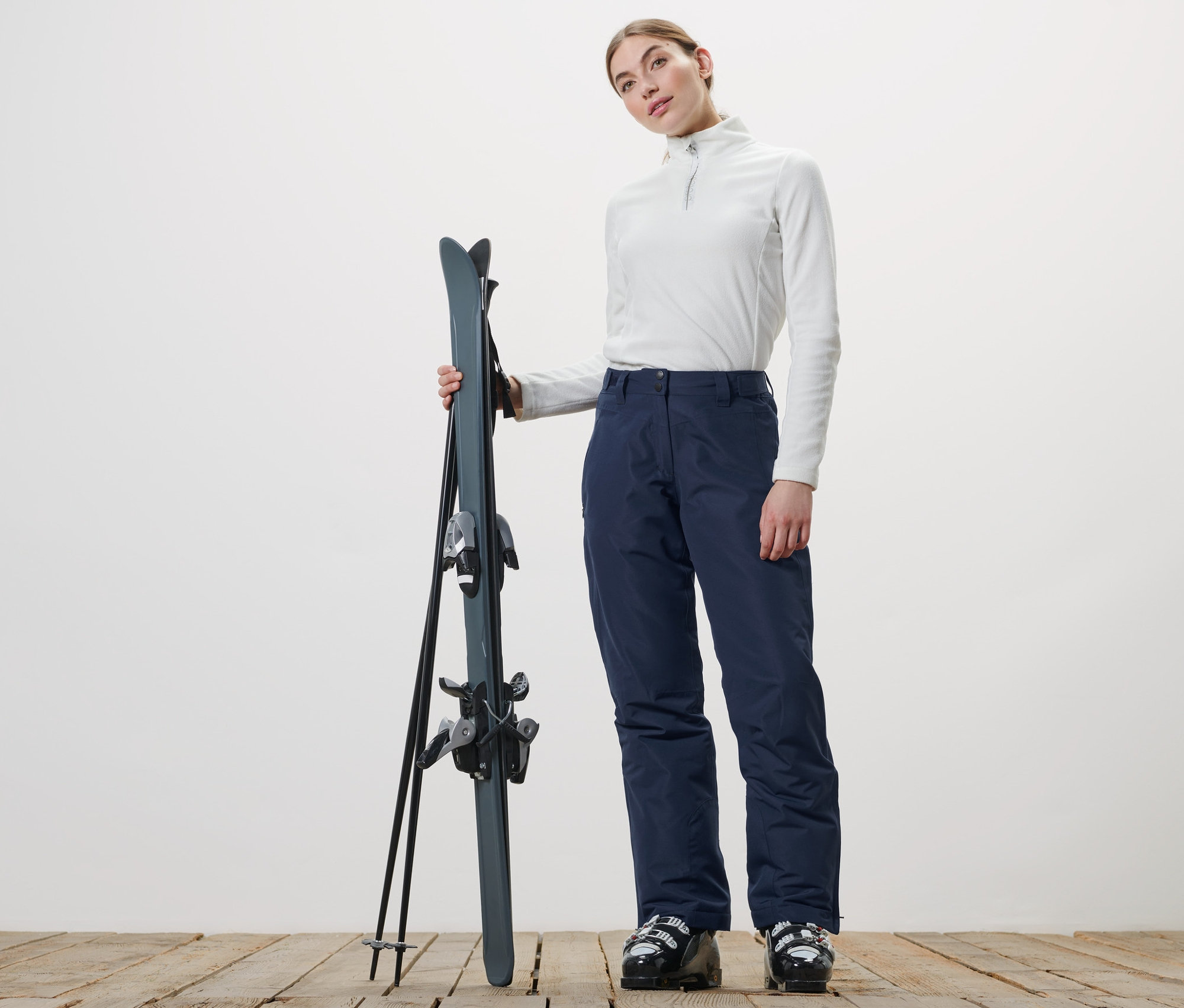 Žena drží lyže a má na sobě lyžařské kalhoty.