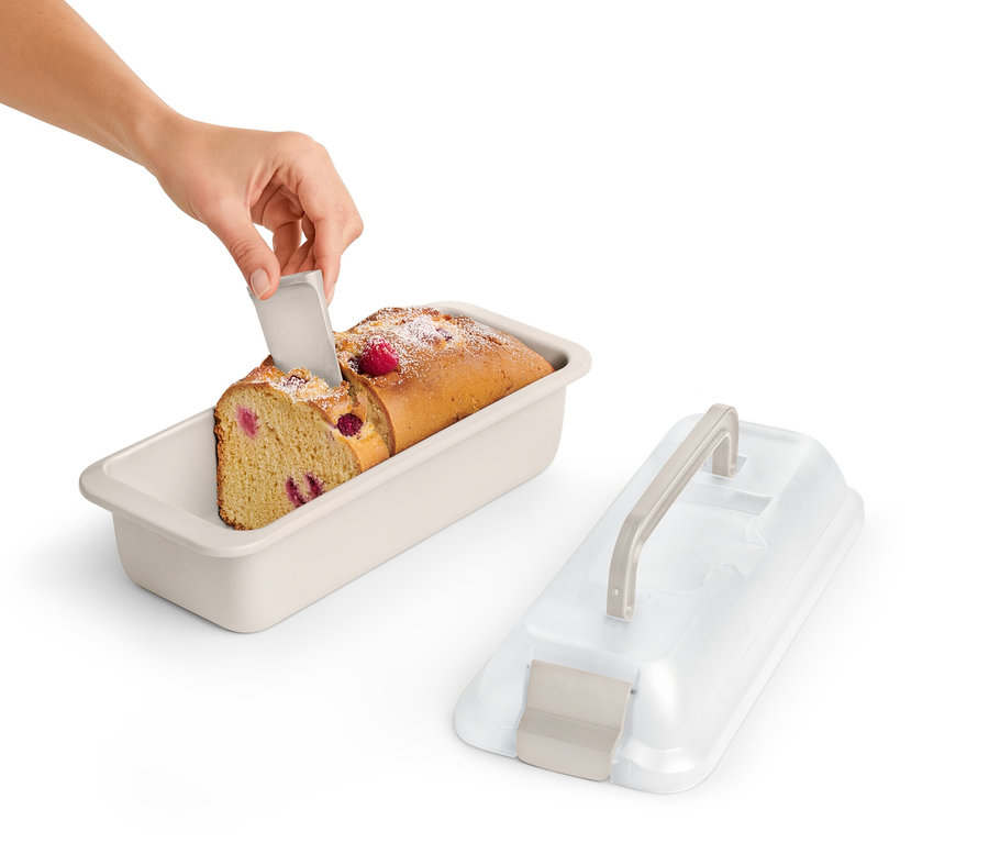 Ruka krájí kousek malinového koláče v hranaté formě s přepravním víkem.