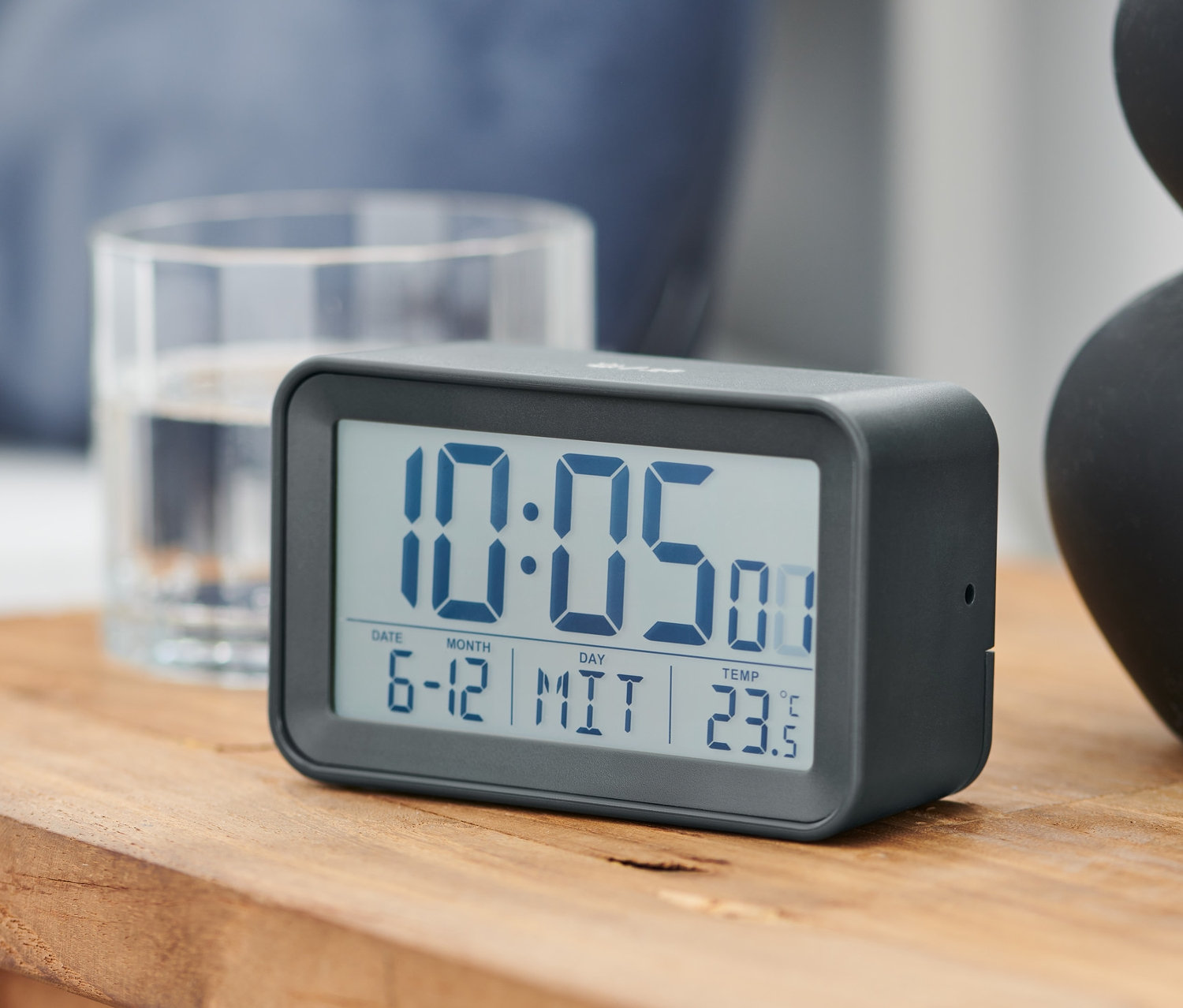 Šedý rádiový LCD budík na dřevěném stolku ukazuje čas 10:05.