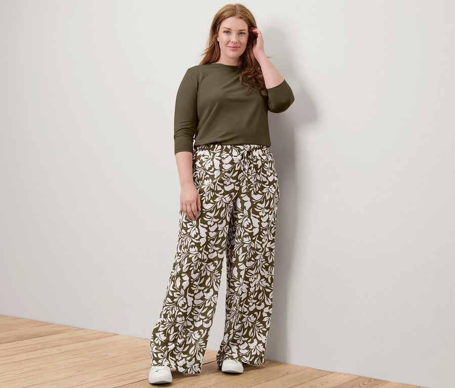 Žena pózuje v khaki tričku s tříčtvrtečním rukávem a vzorovaných kalhotách s gumou v pase.