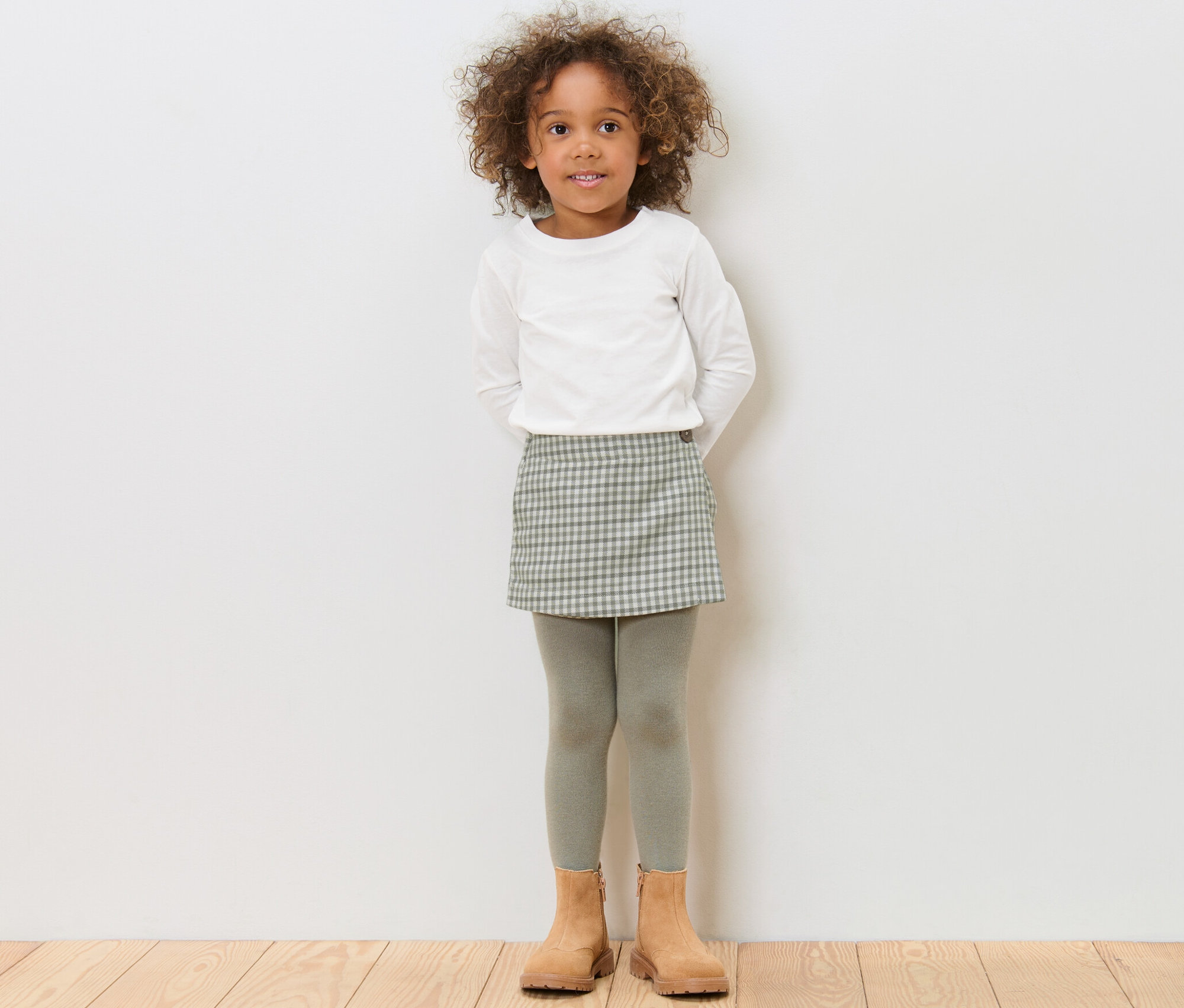 Dívka stojí a má na sobě dívčí kalhotovou sukni, bílé tričko a hnědé boty.