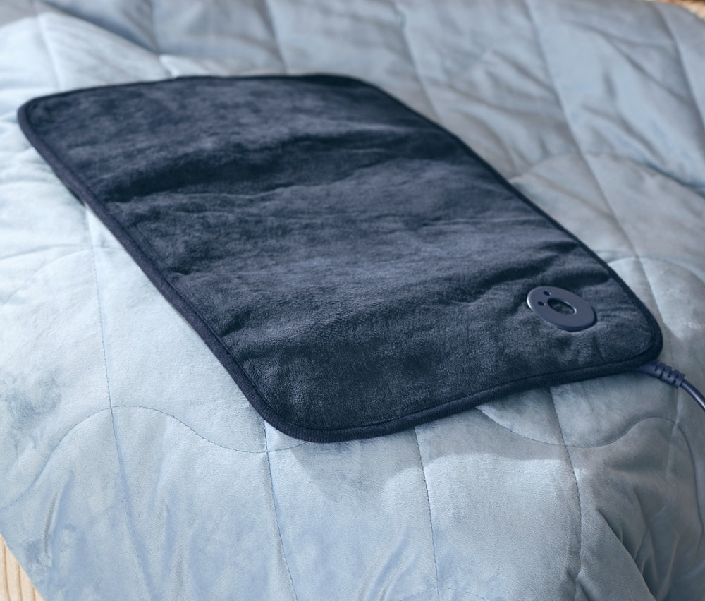 Modré vyhřívané boty a malá elektrická vyhřívací poduška leží na relaxační zátěžové dece, 6 kg.