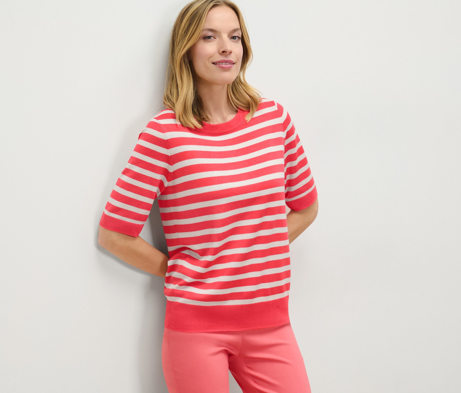 Žena opřená o stěnu má na sobě pruhovaný svetr z jemného úpletu a růžové kalhoty.