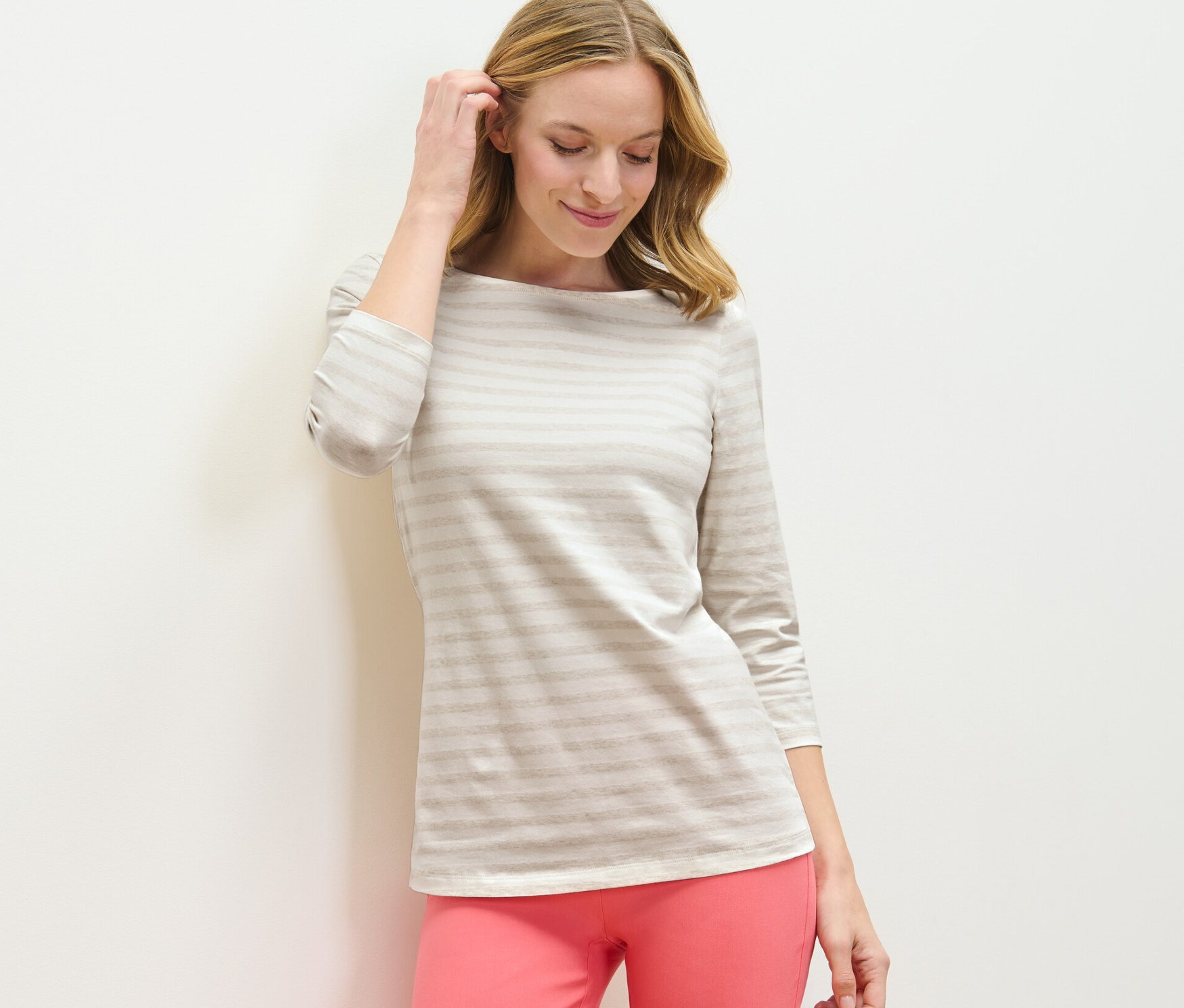 Žena se dotýká vlasů a má na sobě triko s 3/4 rukávem a proužky a růžové kalhoty.
