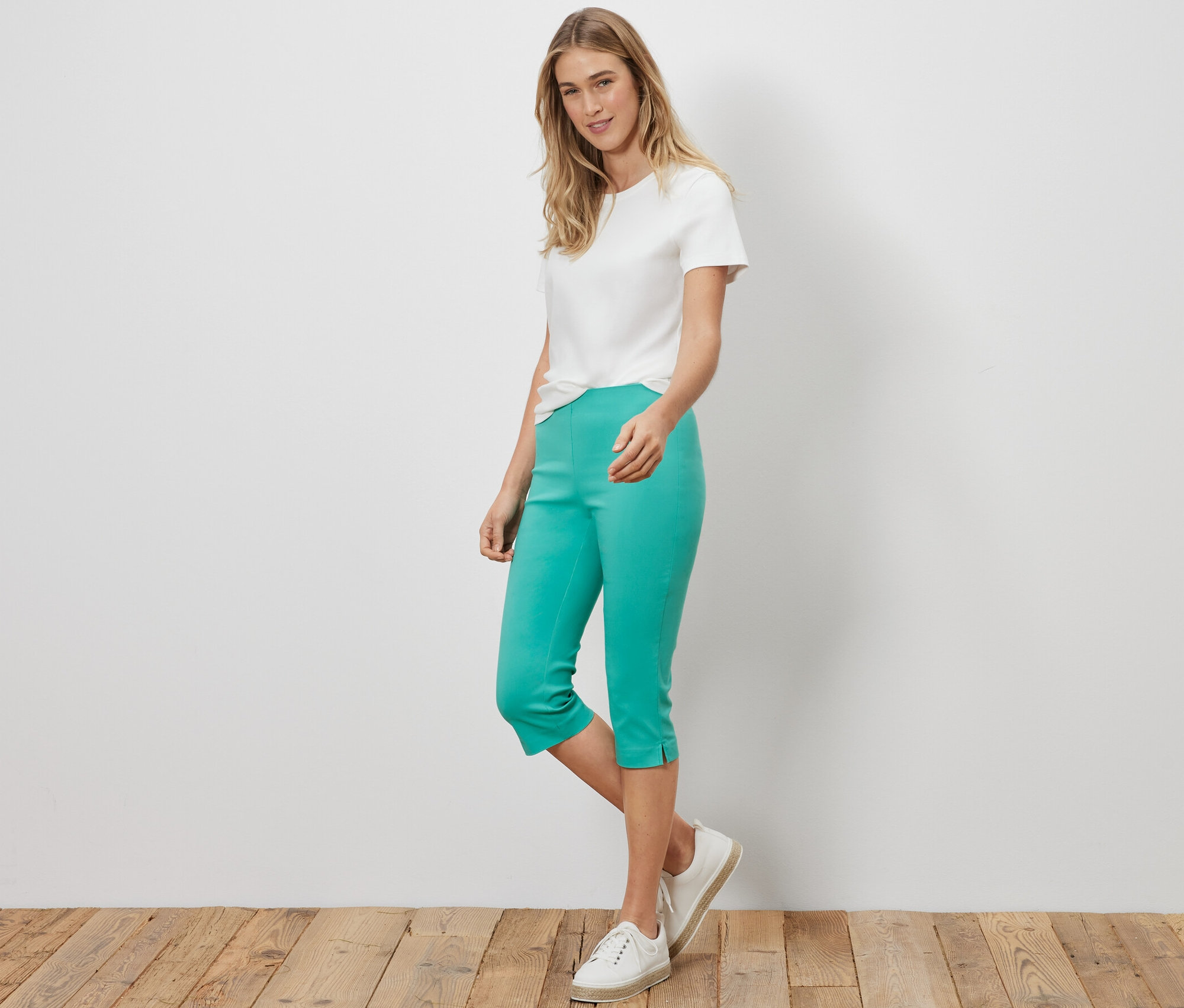 Žena stojí a má na sobě bílé tričko a 3/4 tyrkysové strečové kalhoty.