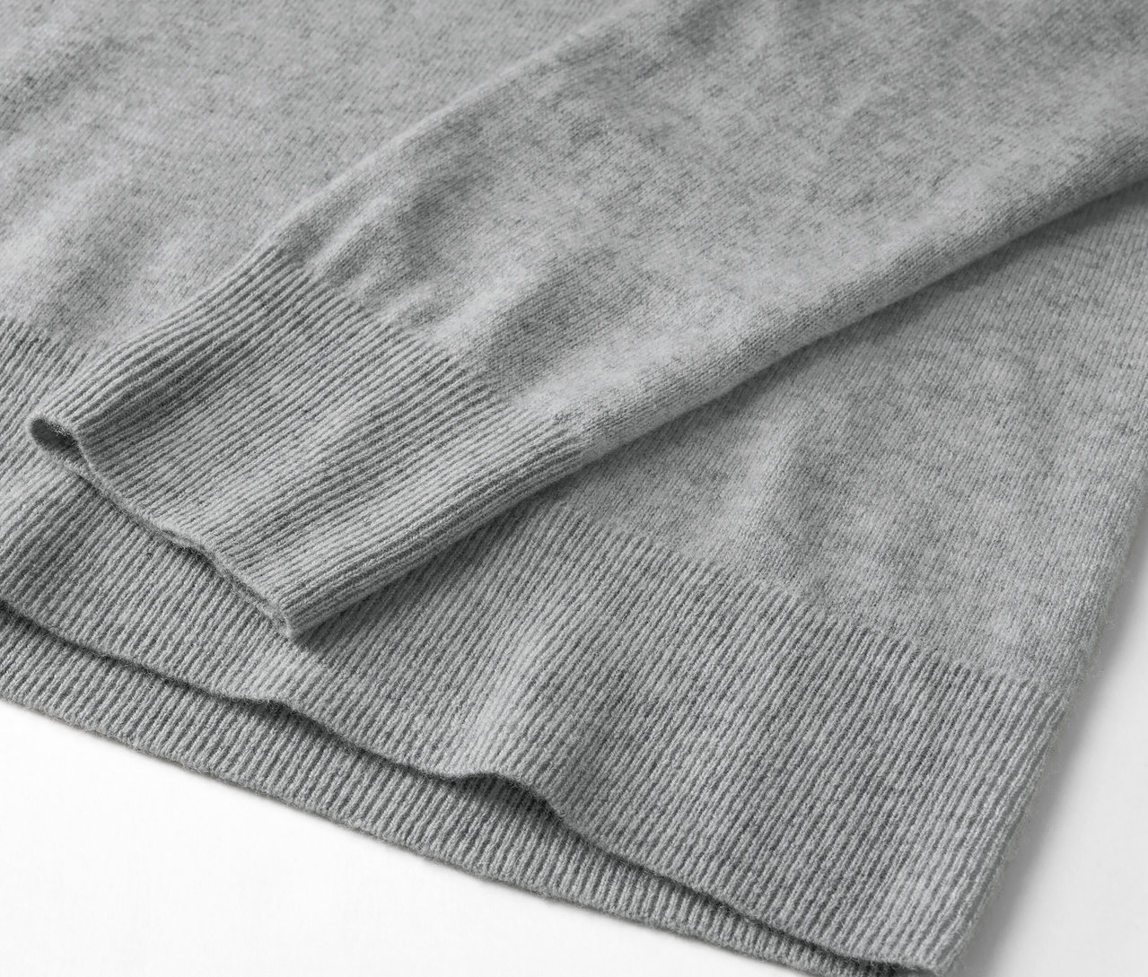 Detail šedého svetru s dlouhým rukávem a žebrovaným lemem.