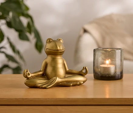 Zlatá soška žáby sedí v meditační pozici vedle skleněné nádoby se zapálenou svíčkou.
