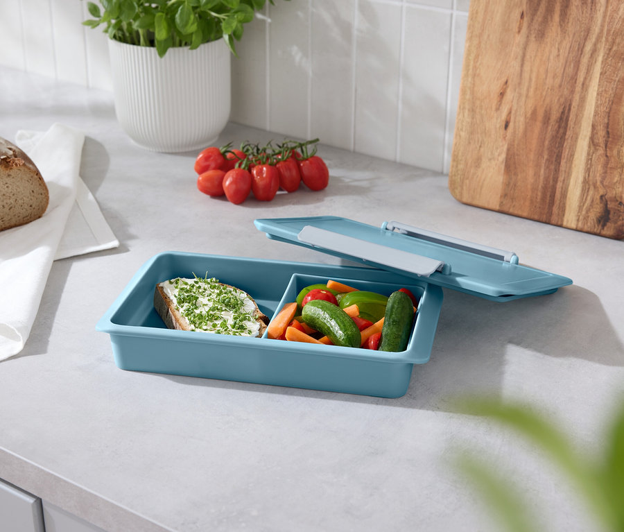 Chléb, rajčata, bazalka a modrý box na svačinu s otevřeným víkem na lince.