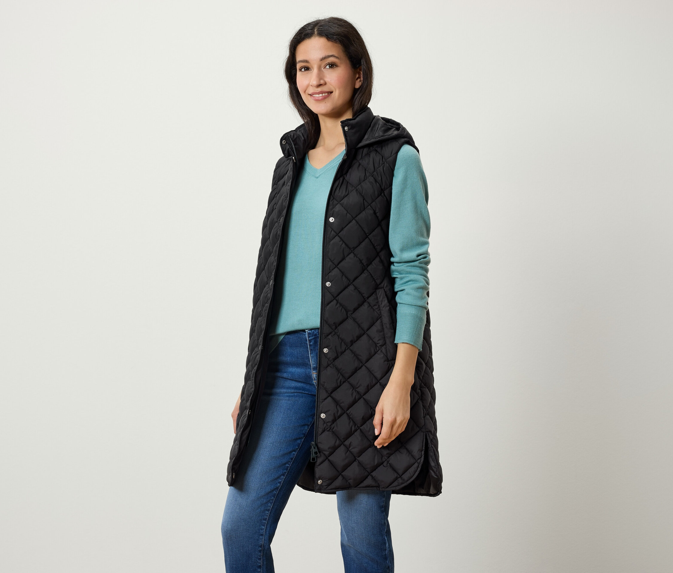 Žena má na sobě dlouhou černou prošívanou vestu s kapucí, modrý svetr a džíny.