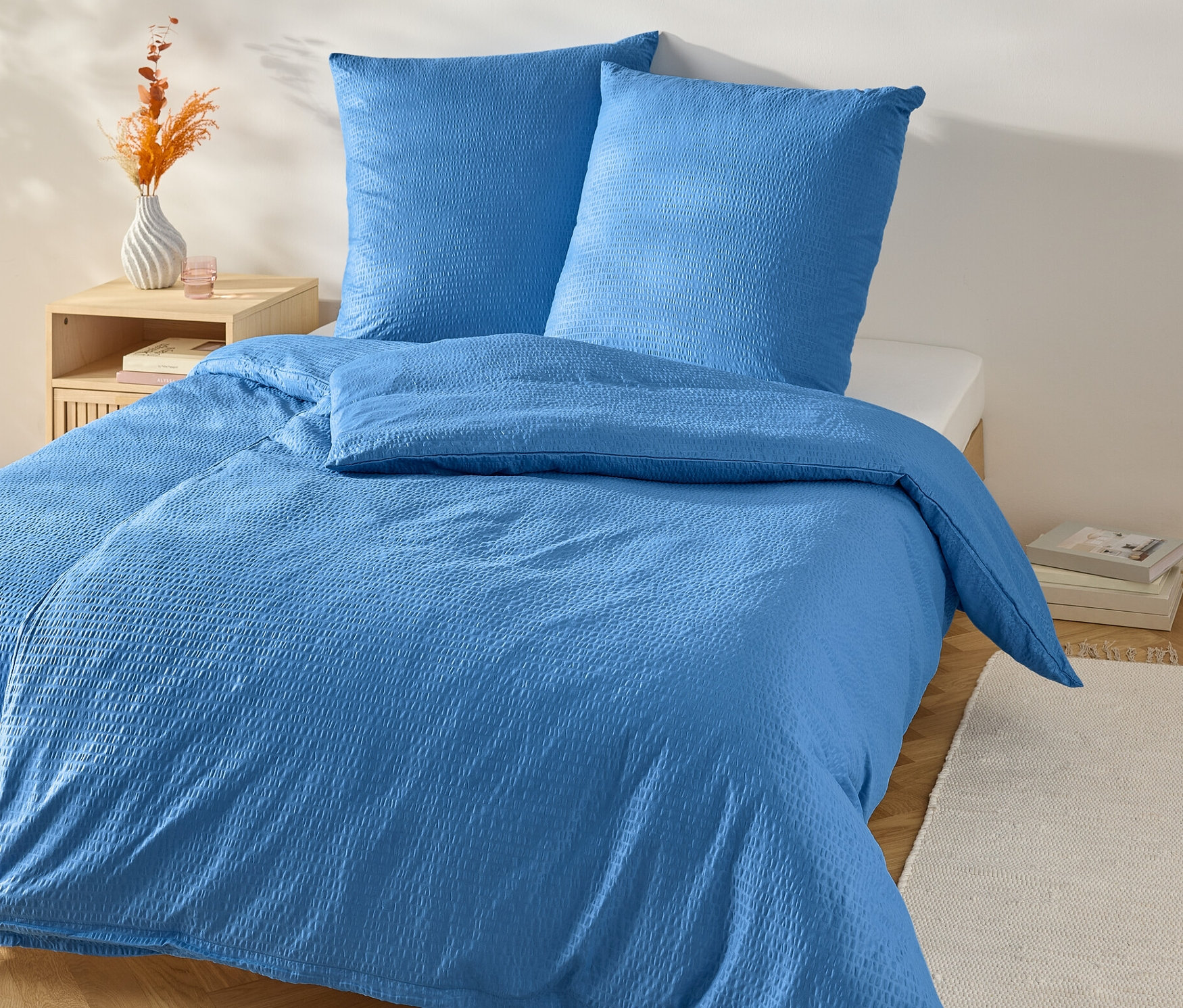 Modrá krepová ložní souprava standardní velikosti na posteli s nočním stolkem.