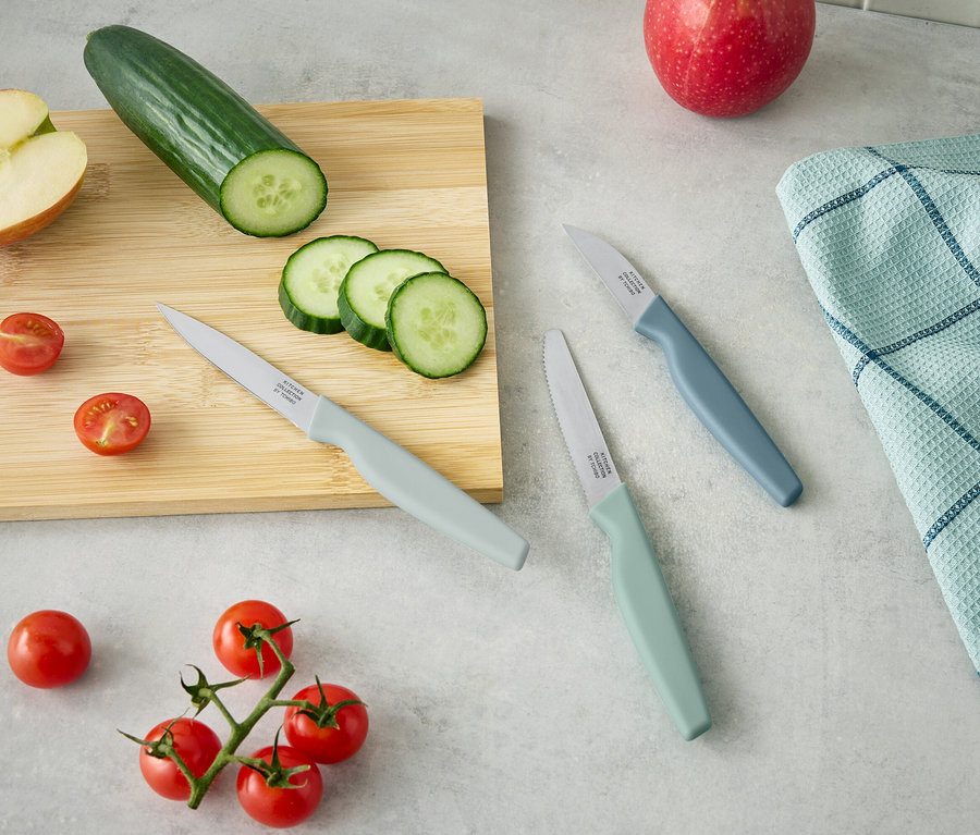 Na dřevěném prkénku leží půlka jablka, okurka a nakrájené plátky okurky. Vedle leží tři nože a cherry rajčata.