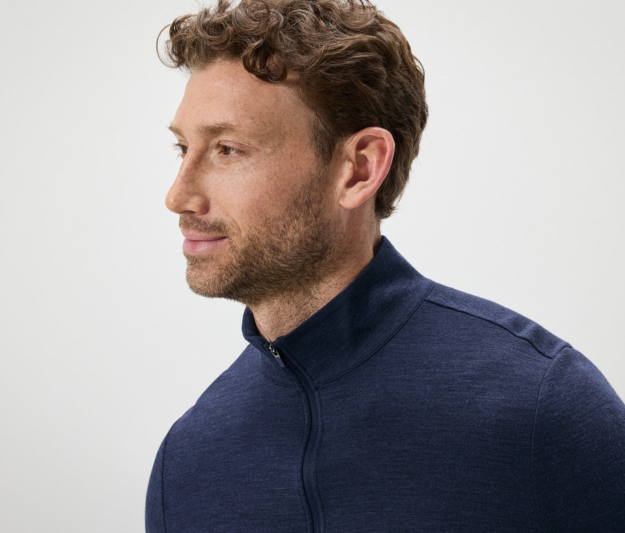 Portrét muže s hnědými vlnitými vlasy a vousy, oblečeného do modrého svetru na zip.