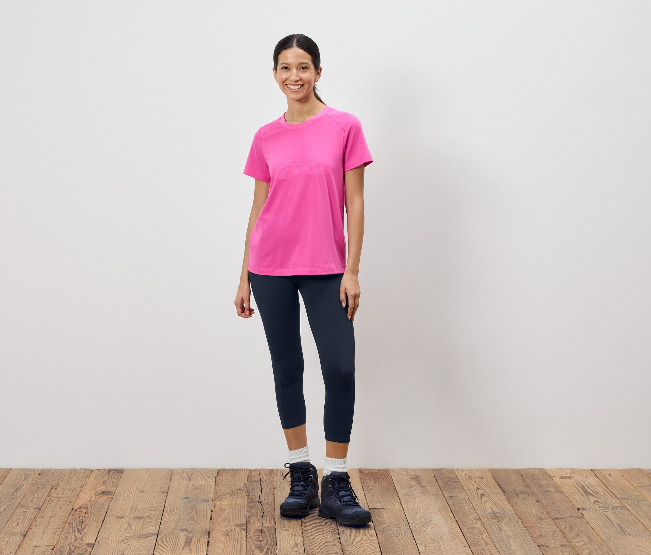 Žena stojí před bílou stěnou a má na sobě růžové tričko, tmavé legíny a trekové boty.