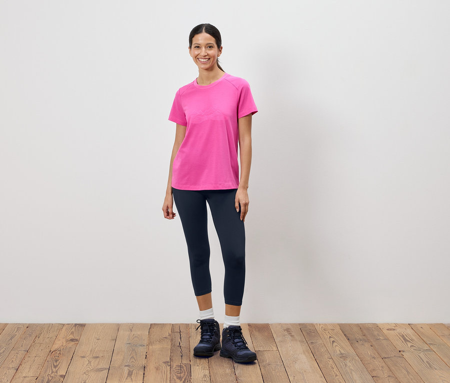 Žena stojí před bílou stěnou a má na sobě růžové tričko, tmavé legíny a trekové boty.