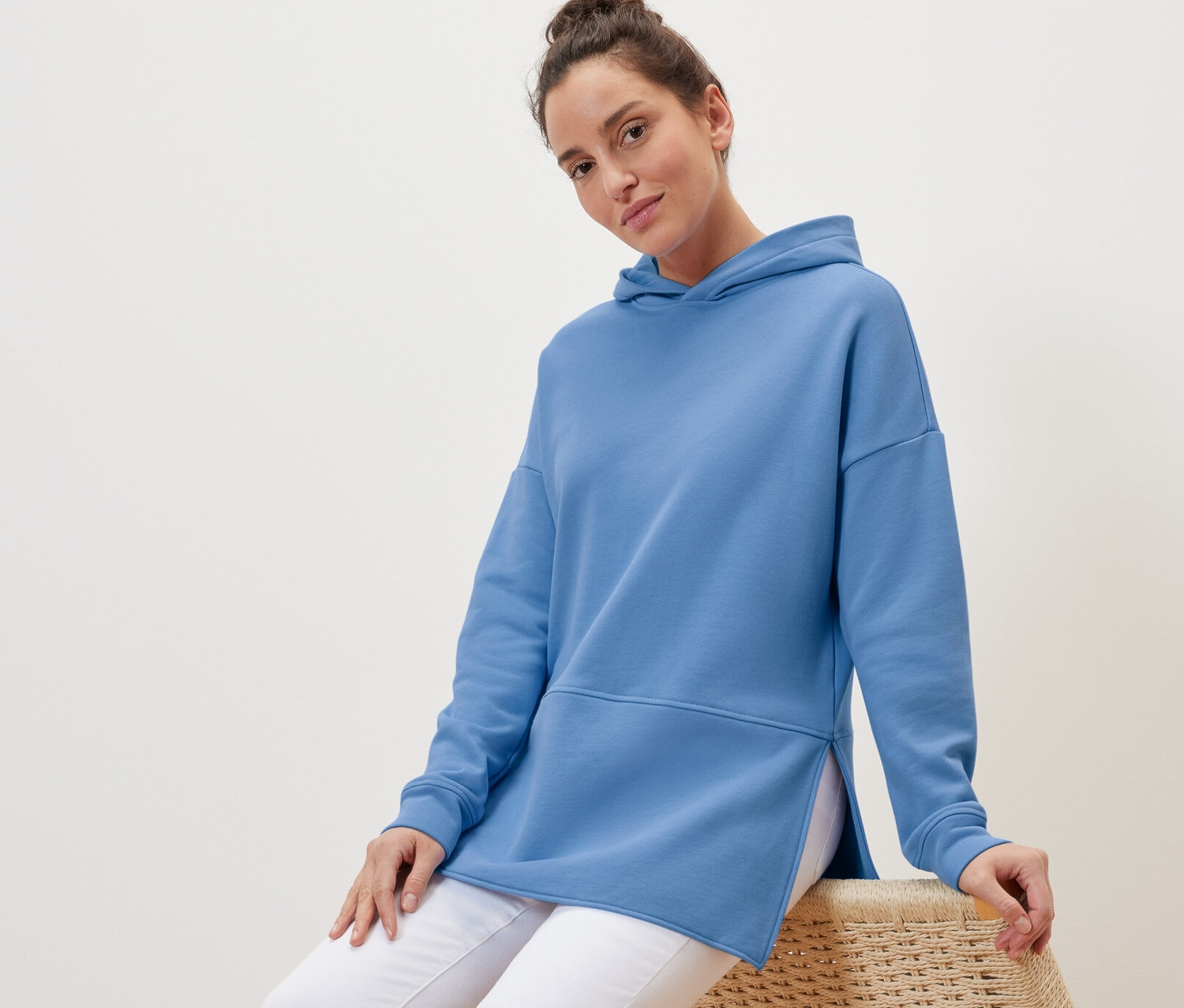 Žena sedí na proutěném křesle a má na sobě světle modrou dlouhou mikinu s kapucí.