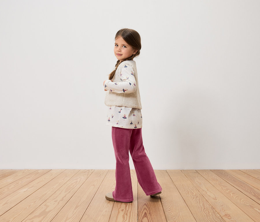 Dívka s hnědými vlasy a copánky pózuje v topu s potiskem, vestě, vínových kalhotách a botách.