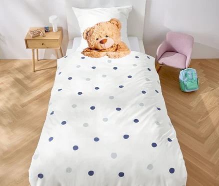 V pokoji stojí postel s bílým povlečením s modrými puntíky a plyšovým medvídkem. Nad postelí visí girlanda z fialových trojúhelníků.