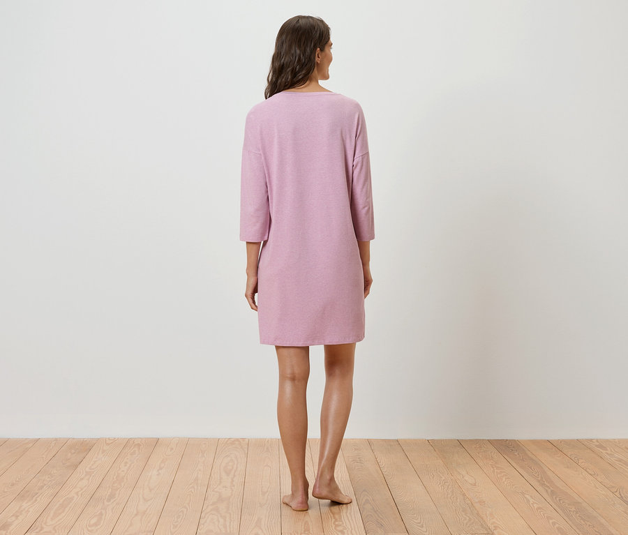 Žena stojí zády k fotoaparátu a má na sobě růžové šaty s tříčtvrtečními rukávy.