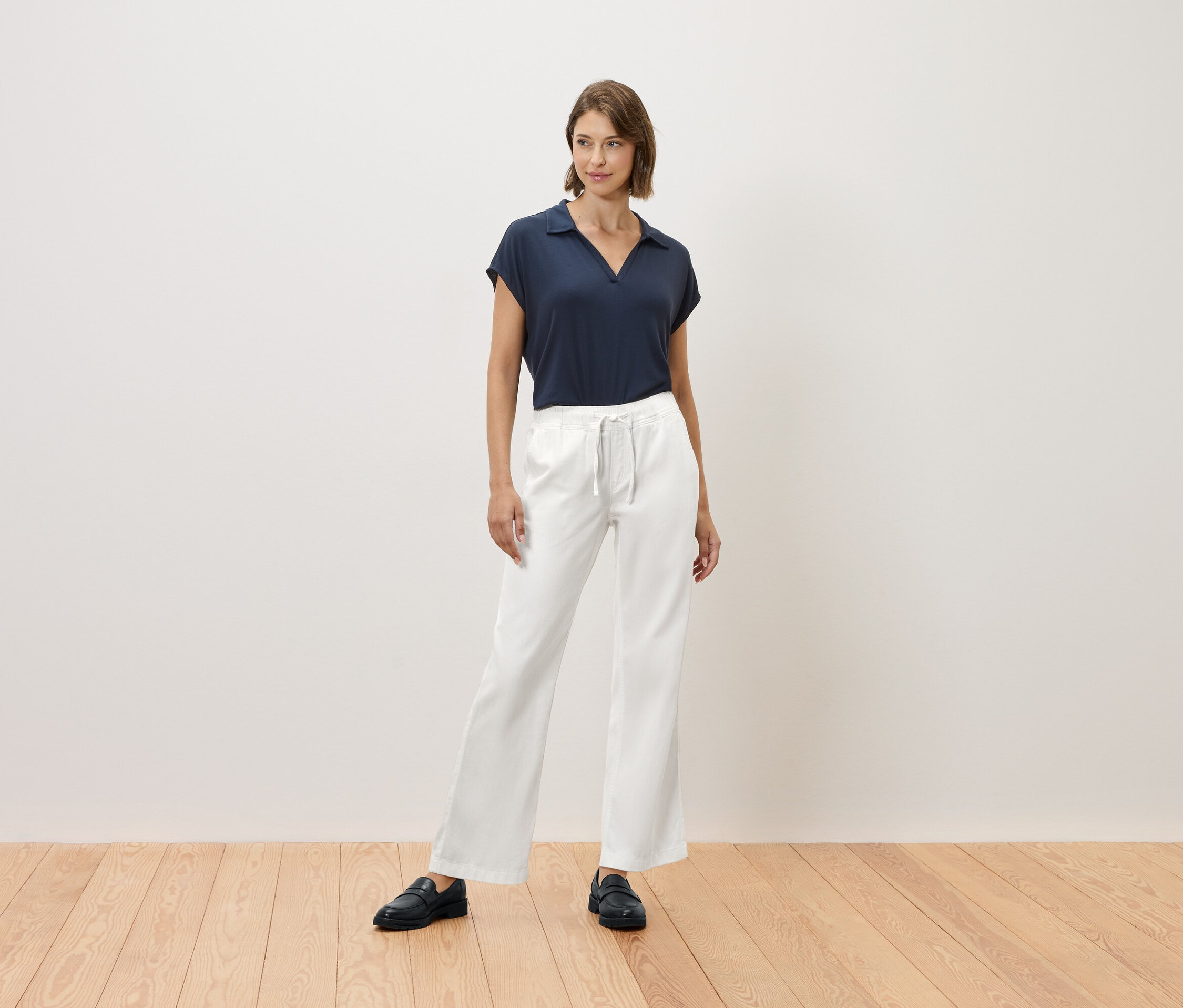 Žena stojí na dřevěné podlaze a má na sobě tmavě modré tričko a bílé kalhoty.