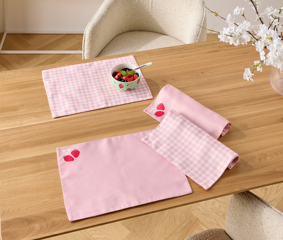 Na dřevěném stole leží růžové prostírání, jedno s jahodovým vzorem, miska s bobulemi a váza s kvetoucími větvičkami.