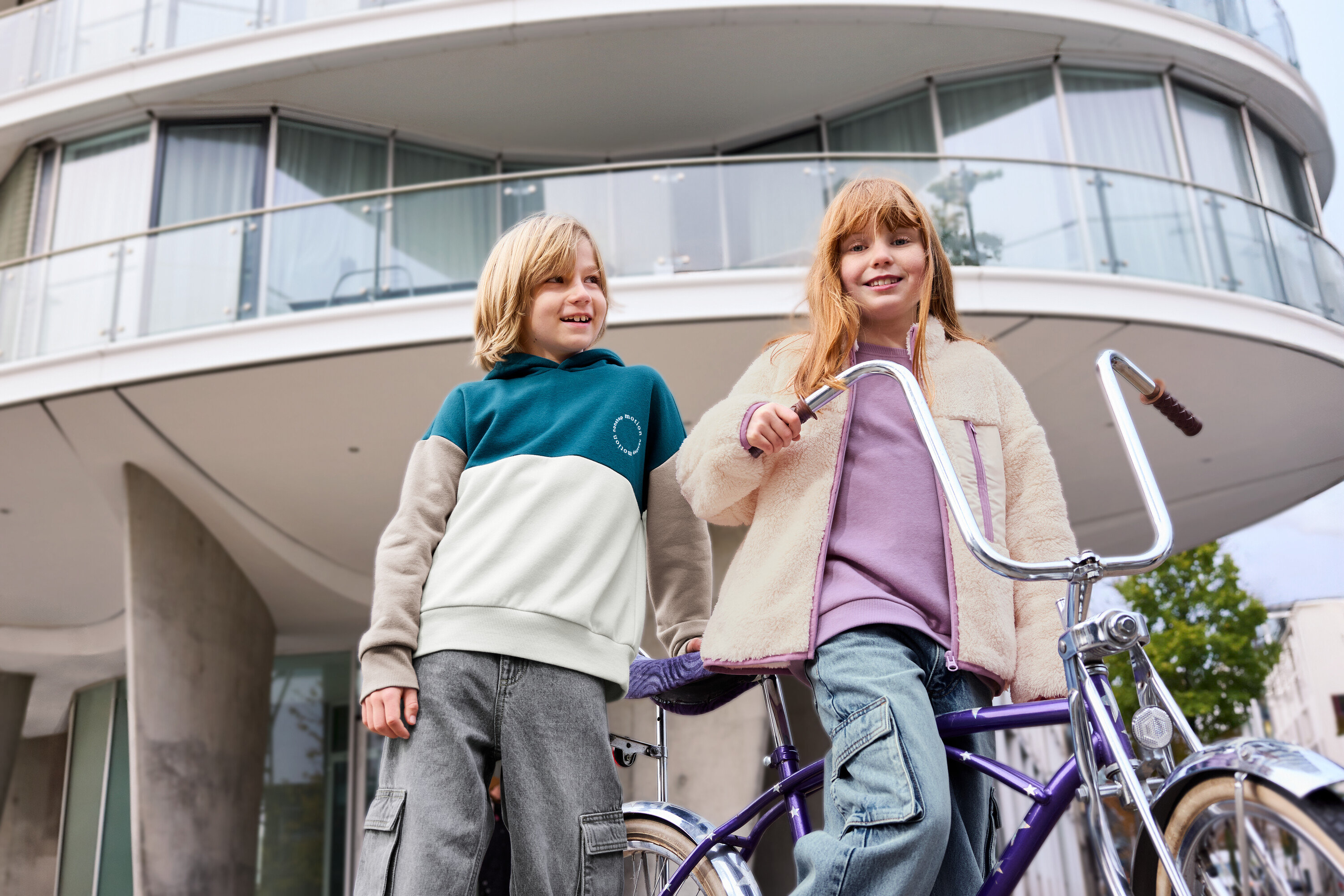 Dvě děti stojí před budovou, jedno dítě má na sobě levandulovou dětskou mikinu s počesanou vnitřní stranou.