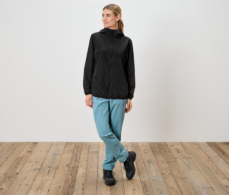 Žena v černé nepromokavé bundě s kapucí, světle modrých kalhotách a černých botách stojí před bílou stěnou.