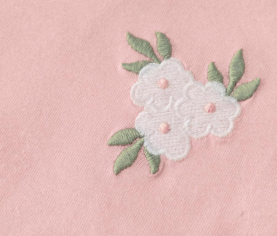 Detail tří vyšívaných bílých květů se zelenými listy na růžové látce.