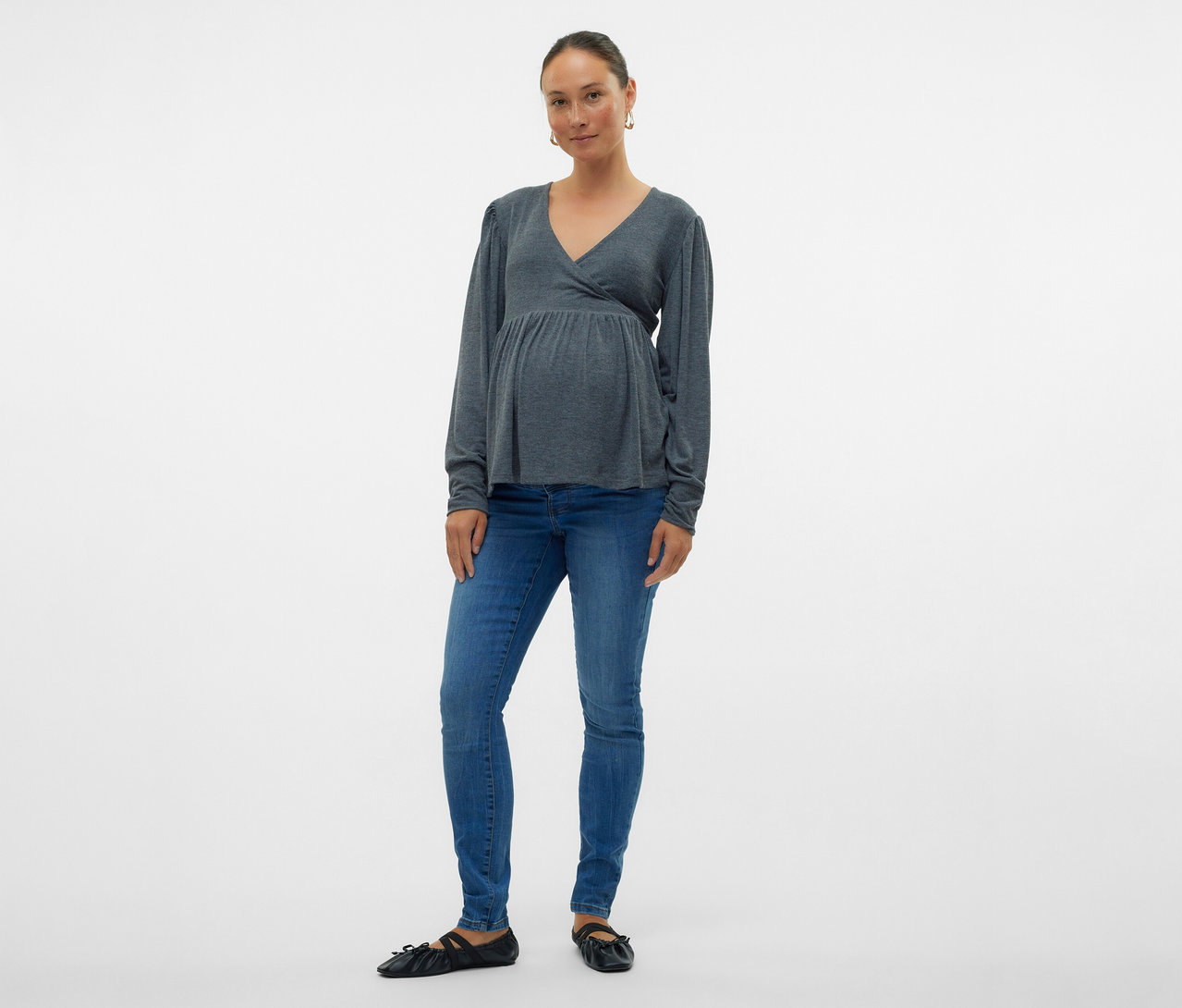 Žena v šedé zavinovací halence a skinny džínách MAMALICIOUS »MLMILA« pózuje na bílém pozadí.