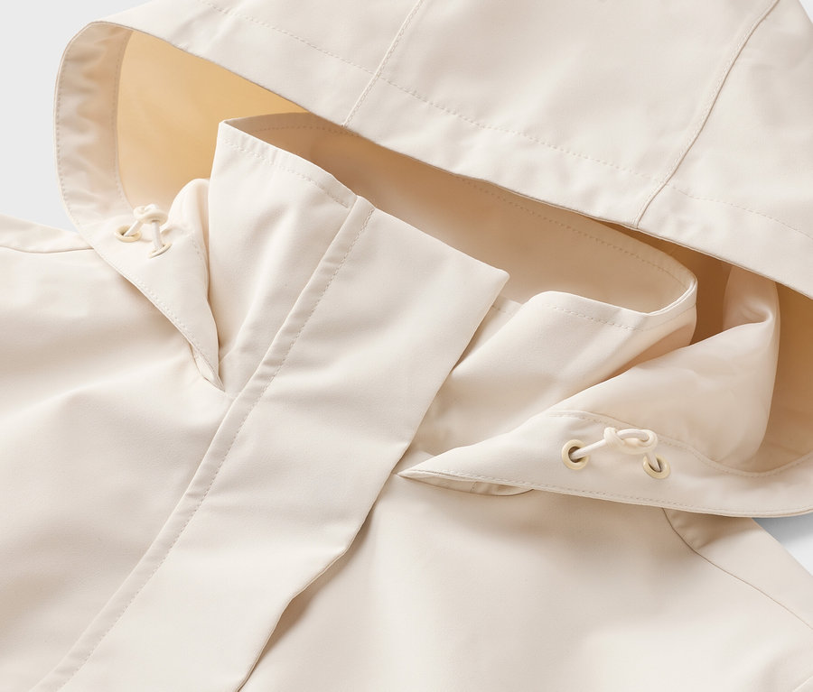 Detailní záběr na světle béžovou bundu do deště s kapucí. Kapuce má světle žlutou vnitřní látku a stahovací šňůrky.