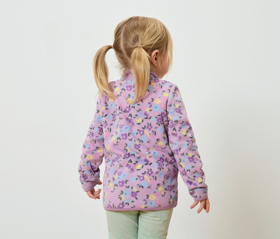 Pohled zezadu na malou dívku s blond copánky, oblečenou do lila fleecové bundy s květinovým vzorem a zelených kalhot.