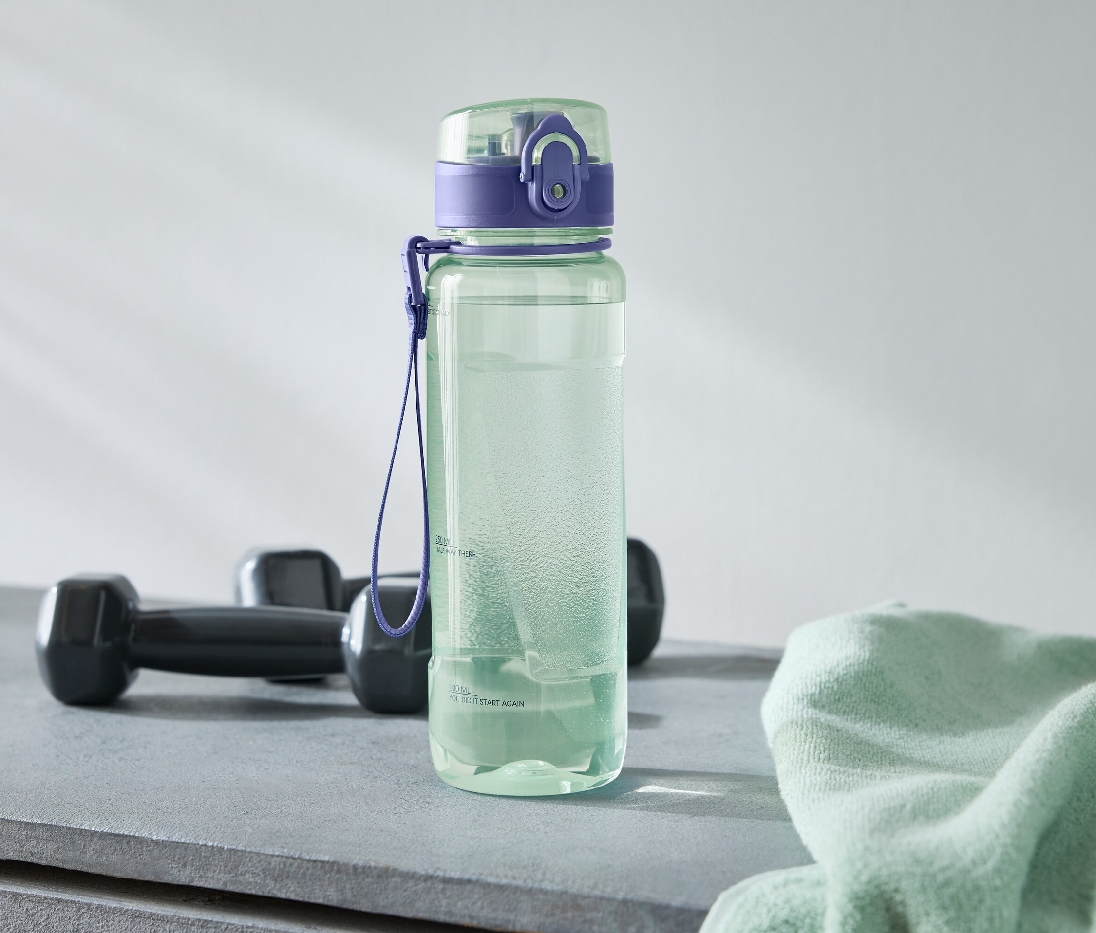 Zelená láhev na vodu s fialovým víčkem stojí vedle dvou černých činek a světle zeleného ručníku na šedém povrchu.