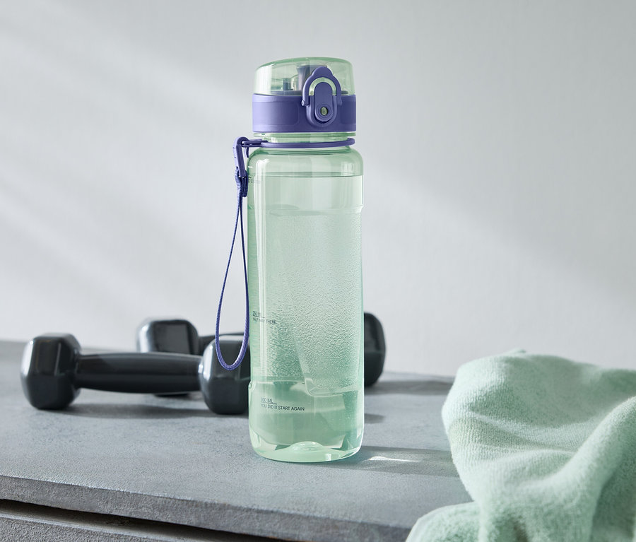 Zelená láhev na vodu s fialovým víčkem stojí vedle dvou černých činek a světle zeleného ručníku na šedém povrchu.