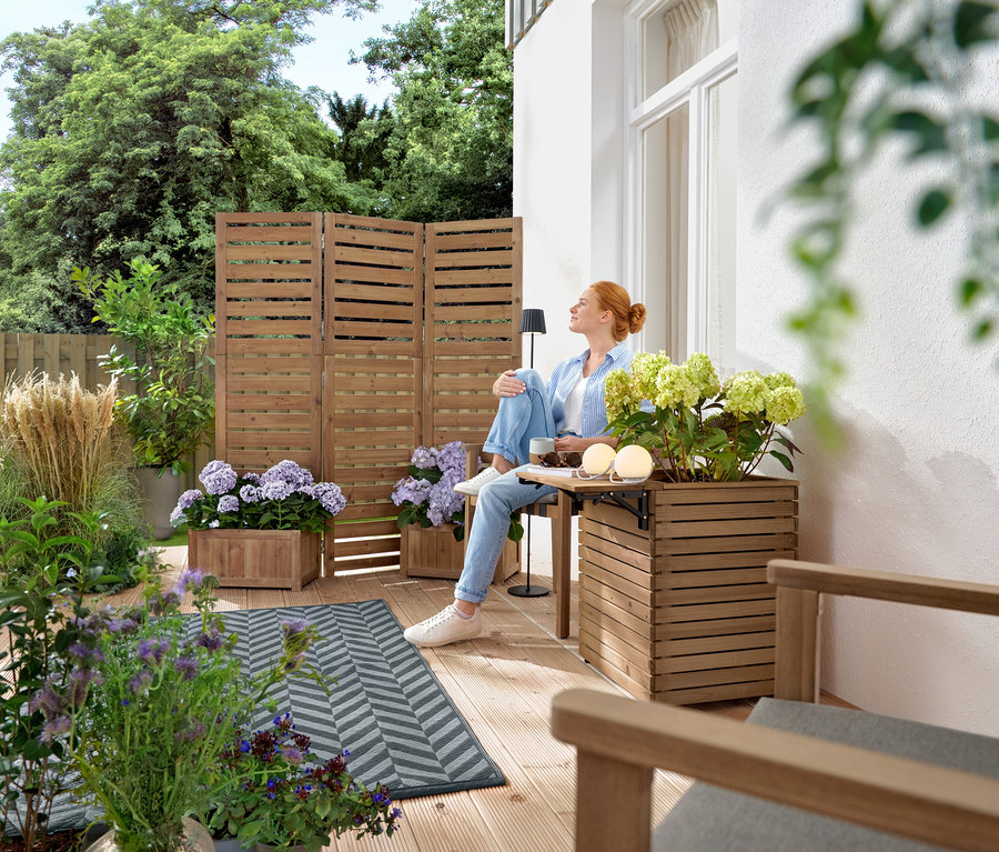 Žena sedí na dřevěné terase se stolem a nápojem, obklopená rostlinami v dřevěných bednách.