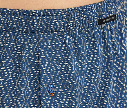 Detail modrých žerzejových boxerek SCHIESSER s bílým kosočtvercovým vzorem, elastickým pasem, černým štítkem SCHIESSER a modrým knoflíkem s oranžovým prošíváním.