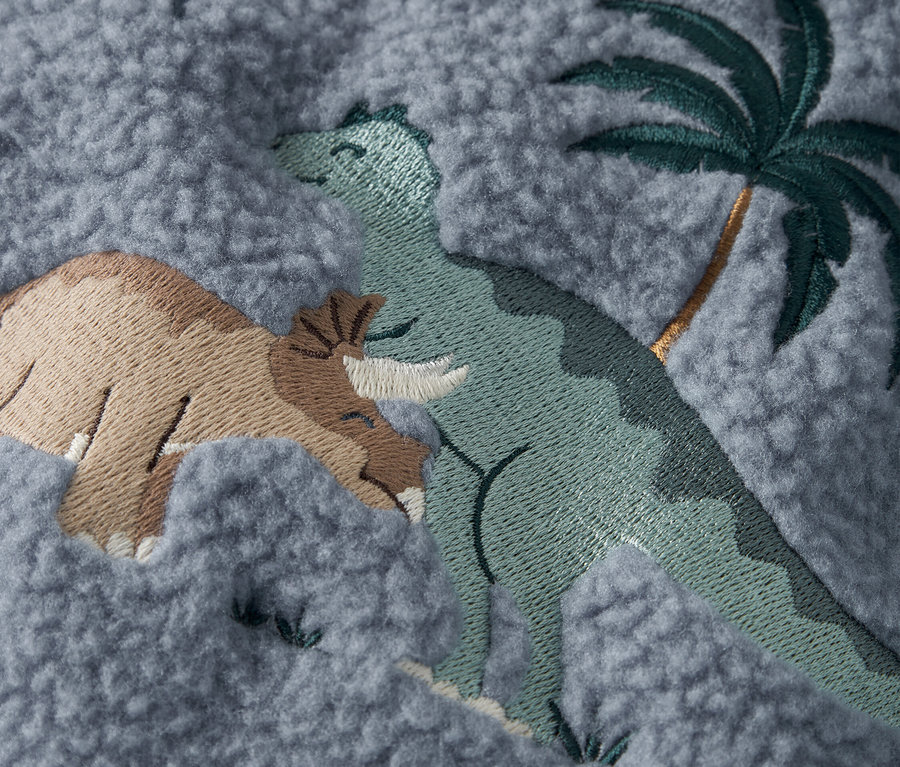 Detail šedé látky s vyšitými dinosaury a palmou.