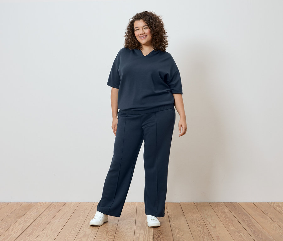 Žena s kudrnatými vlasy v tmavě modrém tričku a kalhotách před bílou stěnou.