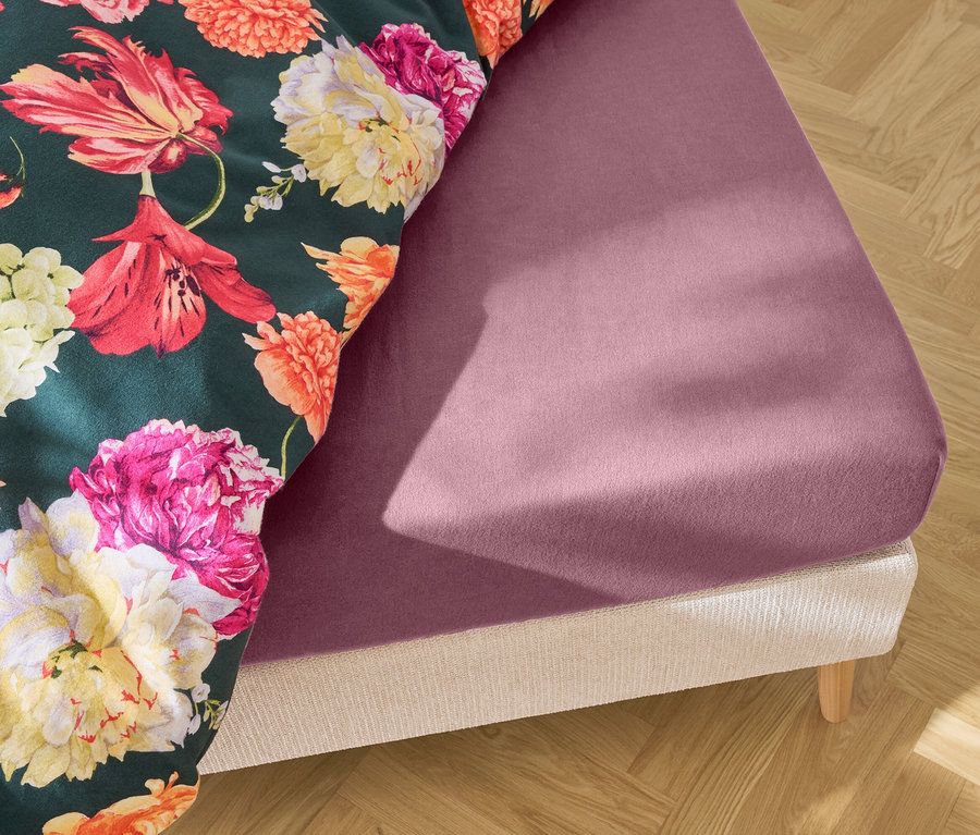 Detailní záběr postele s fialovým prostěradlem a květinovou přikrývkou na dřevěné podlaze.